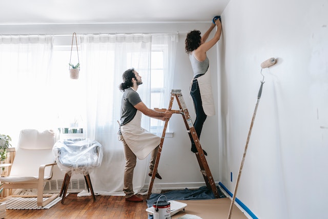 Couple wearing aprons doing house renovations. One person stands on a ladder, noting something on the upper corner of a white wall, the other person holds the ladder. Light comes in through the windows behind them, and their furniture is covered in transparent plastic. 