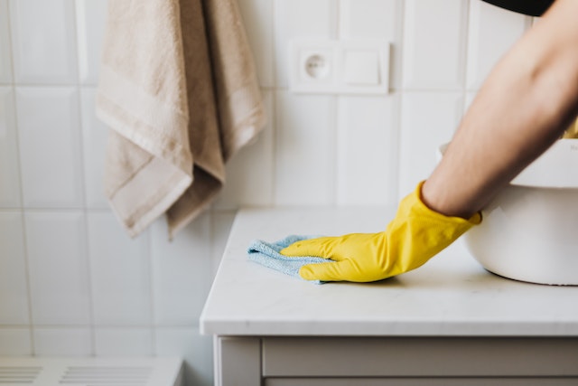 Hand wearing a yellow rubber glove wipes down a white bathroom counter using a light blue washcloth. 