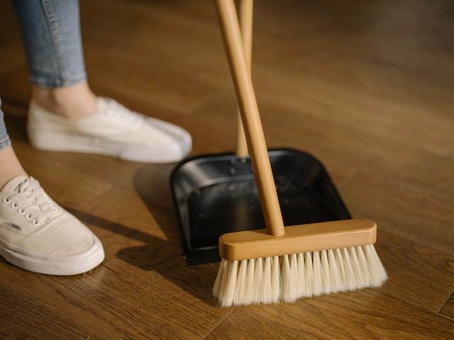 Person wearing light blue jeans and white sneakers standing beside brown wooden broom and a black dustpan