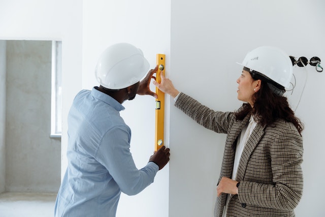 Two people wearing white hard hats holding a level against a wall
